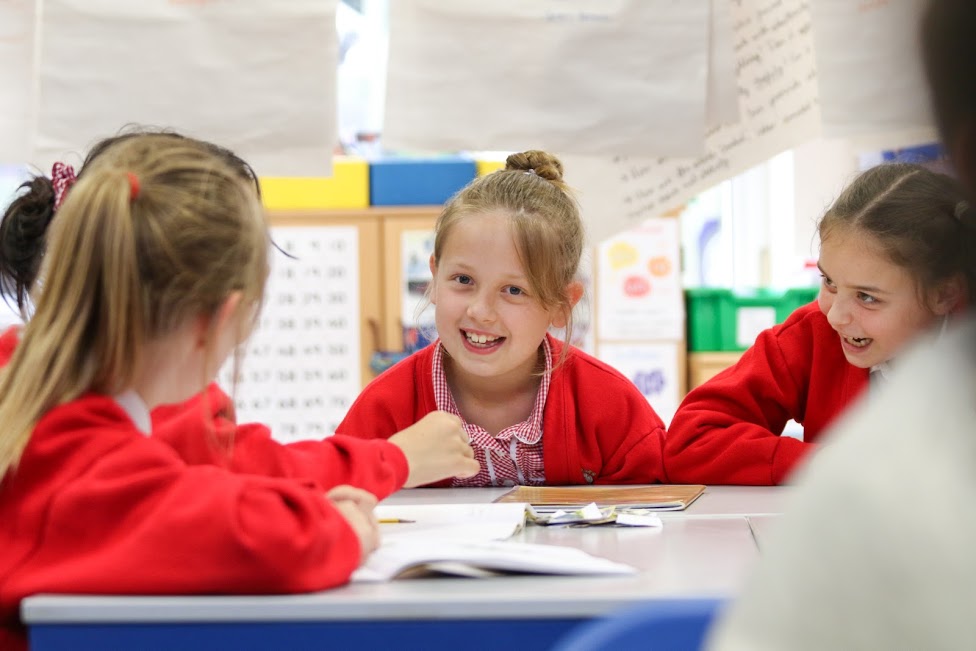A group of four female pupils are seen sat around a table together and smiling at one another. One of the girls is seen looking directly at the camera.