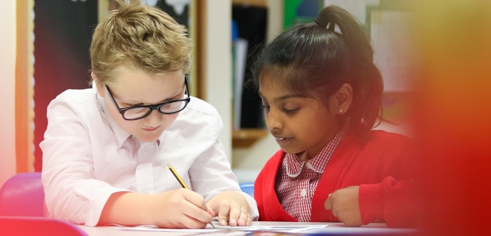 Two pupils, a boy and a girl, are photographed sitting next to one another at a desk, concentrating hard on their work. The boy is seen writing with a pencil in his right hand.